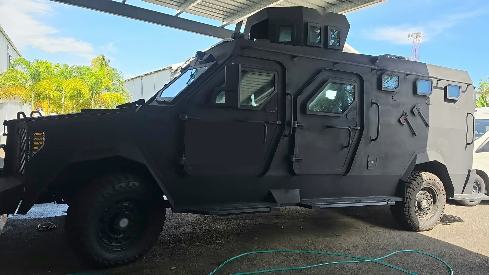 Vehículo táctico blindado de gran formato visto de lado, al aire libre junto a una nave industrial